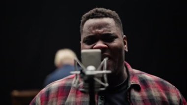 Bir stüdyoda siyah bir fon eşliğinde duygusal bir şarkı söylerken bir adamın yüz ifadelerini gösteren yakın çekim.