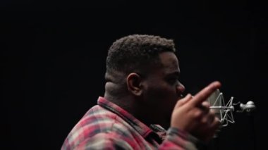 Ekose gömlek giyen bir adam stüdyoda şarkı söylerken parmaklarını işaret eder.