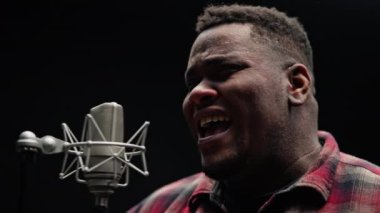 Stüdyoda gümüş bir mikrofona tutkuyla şarkı söyleyen bir adam yoğun bir duygu sergiliyor.