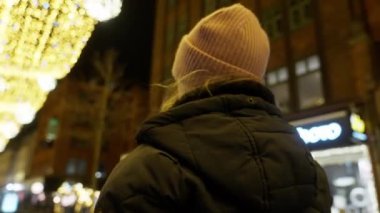 Alçak açılı arka plan, soldan sağa hareket eden pembe bereli ve kışlık ceketli bir kızın Noel ışıklarıyla yürüdüğünü gösteriyor.