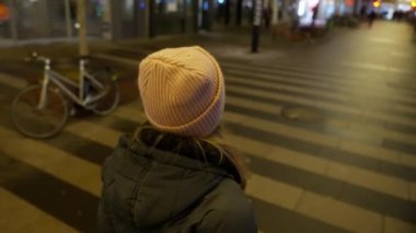 Arka planda pembe bereli ve gri ceketli bir kız var. Kaldırımda bulanık bir zeminde yürüyor.