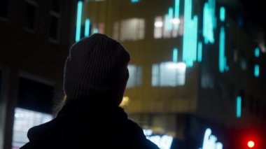 Gece vakti mavi ışıkları olan bir binanın önünde duran bir kızın düşük açılı görüntüsü.