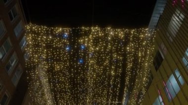 Aşağıdan çekilen bir video binalar ve karanlık gece gökyüzü arasında asılı duran sicim ışıkları ve parlak küreleri gösteriyor.