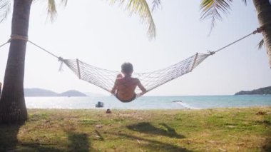 Görüntüyü büyüt, Tayland 'da gündüz vakti deniz kenarında sallanan bir çocuğun arka görüntüsünü göster.