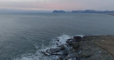 Kayalık kıyı şeridi üzerinde dron, denizin sakin suları, İzlanda 'da gün batımında dağ sırasının uzak bir görüntüsü.