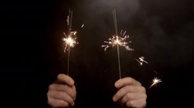 İki çift yanan ve dumanı tüten maytapları koyu bir arka plana doğru kaldıran elleri gösteren bir video.