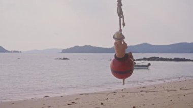 Şamandıra topunda sallanan genç bir çocuğun videosu. Tayland 'da bir plajda gün içinde ileri geri sallanıyor.