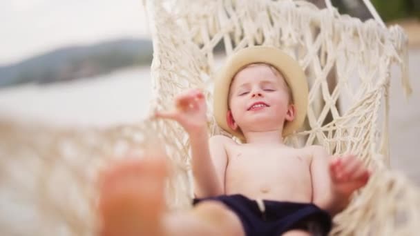帽子をかぶった少年が 昼間のタイのビーチでハンモックに揺られながらカメラの前で微笑む — ストック動画