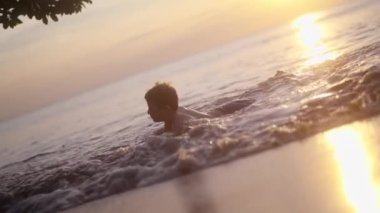 Tayland 'da gün batımında kıyıya vuran dalgalarla plaj suyunda yatan bir çocuğun videosu.