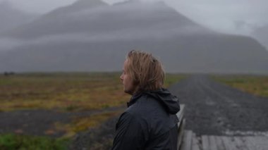 Yağmurluk giyen sarışın bir adamın bulutlu bir günde dağın yanındaki köprüde dikilirken yan görüntüsü.