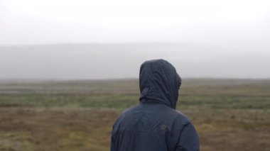 Yağmurluk giymiş bir adamın, yakınlardaki dağ manzaralı ve bulutlu gökyüzü manzaralı bir tarlanın ortasında dikilirken, geriye doğru görüntüsü.