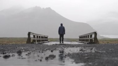 Yağmurluk giyen bir adam, yağmurda dağın yakınındaki bir köprüde duruyor.