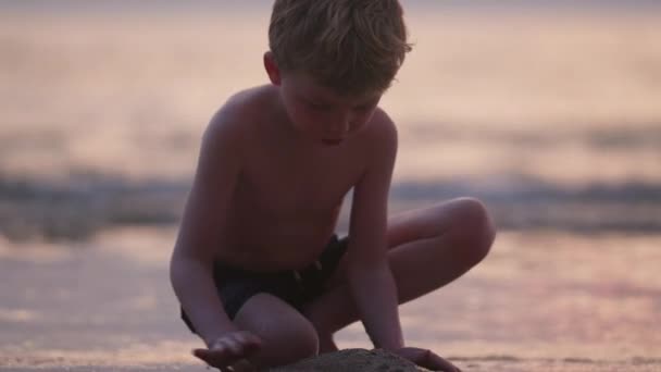 日没の間にタイのビーチの海岸線に沿って砂を積もうとしている少年のビデオ — ストック動画