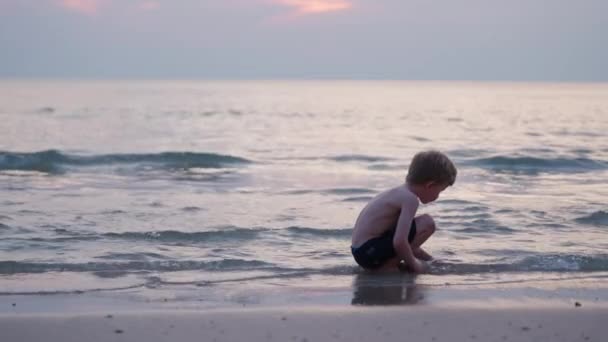 Nagranie Chłopca Kucającego Podczas Zabawy Piaskiem Plaży Podczas Zachodu Słońca — Wideo stockowe