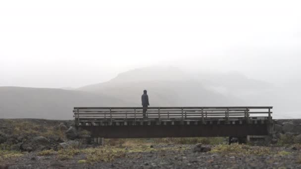 山と雲の空の上に見える小さな橋の上に立ってジャケットに男の広いショットを傾ける — ストック動画