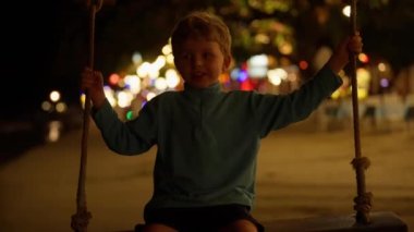 Tayland 'da gece vakti arkasında renkli bokeh ışıkları olan bir plajda salıncakta sallanırken gülümseyen bir çocuğun orta boy fotoğrafı.