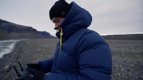 一名身穿夹克的男子和一名贝尼站在冰封的冰岛湖畔看着手中的无人机控制器的侧视图 — 图库视频影像