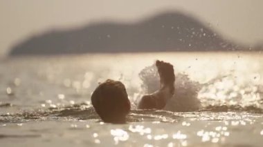 Tayland 'da gün batımında sığ deniz suyunda yatarken ayaklarını çırpan bir çocuğun videosu.