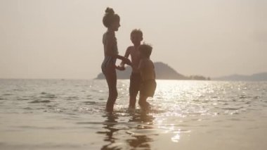 Tayland 'da gün batımında güneş parıltılarıyla sığ sularda atlarken el ele tutuşan çocukların kumsalda eğlendiği bir video.