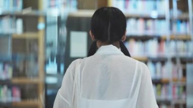 Siyah at kuyruklu ve gözlüklü bir kadının kütüphanenin kitap raflarında ağır çekimde yürüdüğü arka plan görüntüsü.