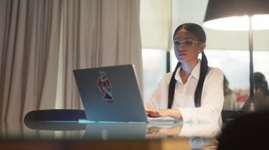 Gözlüklü bir kadın laptopuyla kütüphanede yavaş çekimde yazarken masanın üzerine oturur.