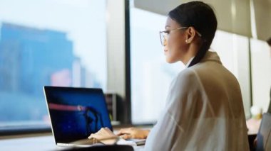 Gözlüklü bir kadının pencerenin yanındaki masada çalışırken çekilmiş bir fotoğraf.