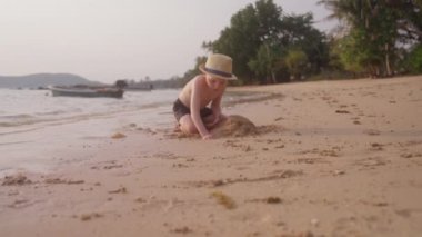 Şapka takıp mayo giyen, Tayland 'da bir kumsalda kum kazan küçük bir çocuğa doğru hareket eden bir video.