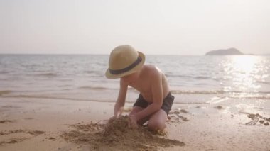 Tayland 'da bir plajda, şapkalı ve yüzme şortlu bir çocuğun gündüz videosu kumu yığılıyor.