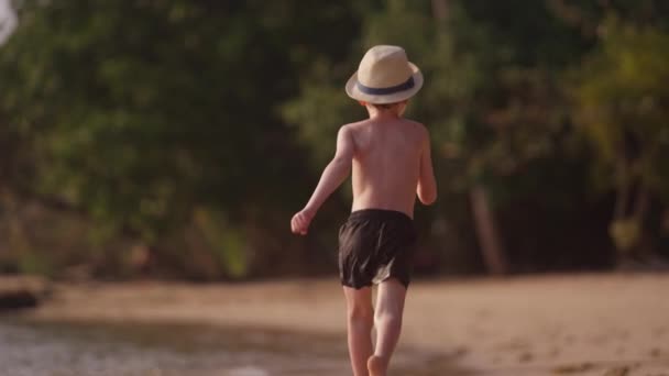 一段录像显示了一个小男孩白天戴着一顶帽子慢吞吞地沿着泰国海滩跑的背景图 — 图库视频影像