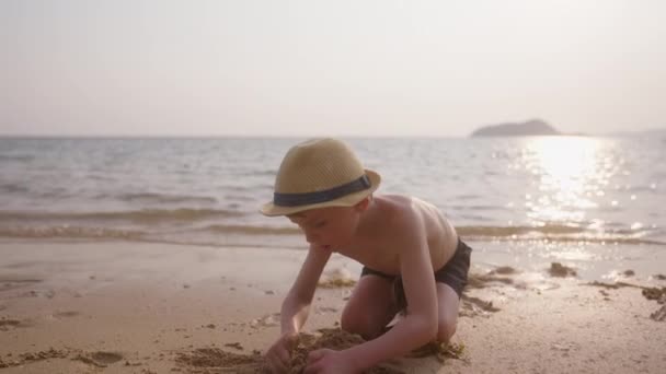 泰国一名男孩白天戴着帽子和游泳裤在海滩边玩沙子的视频 — 图库视频影像