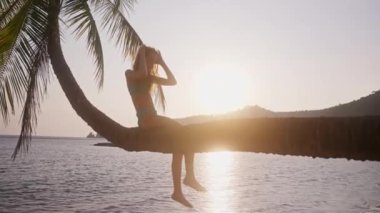 Gün batımında Tayland 'da bir plajda eğilmiş bir palmiye ağacında oturan bir kızın yan görüntüsünü gösteren geniş bir çekim.
