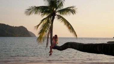 Tayland 'da bir plajda bir palmiye ağacının gövdesinde oturan üç çocuğun yan görüntüsünü gösteren bir video.
