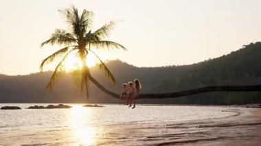 Tayland 'da bir plajda palmiye ağacının gövdesinde oturan üç çocuğun görüntüsü.