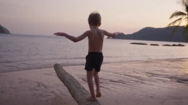 Gün batımında Tayland 'da bir plajda ağaç gövdesinde yürüyen bir çocuğun videosu.