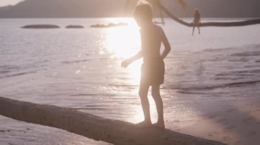 Bir çocuğun ağaç gövdesinde yürüdüğü ve gün batımında Tayland 'da kumlu bir sahilde atladığı bir video.