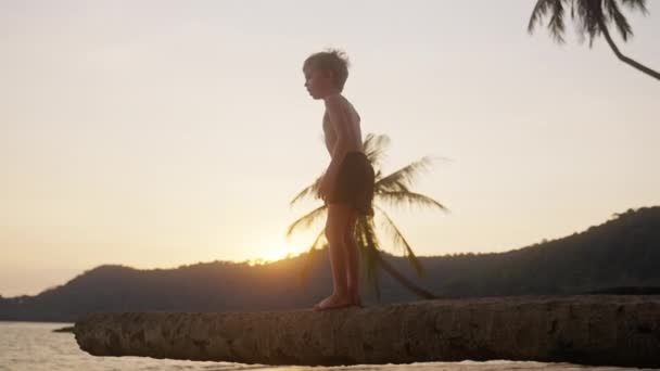 一个男孩在日落时坐在泰国海滩的树干上的视频 — 图库视频影像