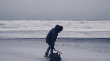 Kışlık ceketli bir adam seyahat çantasını Reynisfjara sahilinin kara kumlarına bırakır ve rüzgârı hissetmek için kollarını kaldırır.