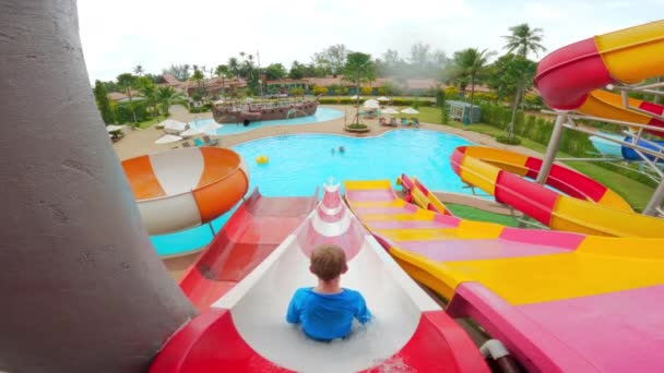 カラフルな水のスライドと タイの昼間の下のプールで滑る少年の背中の景色を示す広いショット — ストック動画