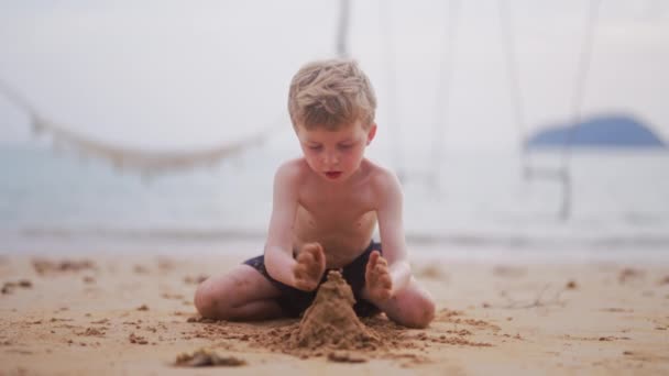 タイで昼間に砂の城を建てるために海岸に砂をゆっくり積んでいる若い男の子 — ストック動画
