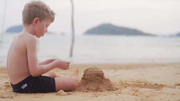 タイの昼間にビーチで建てた砂の山の上に岩を置いた少年の横からの眺め — ストック動画