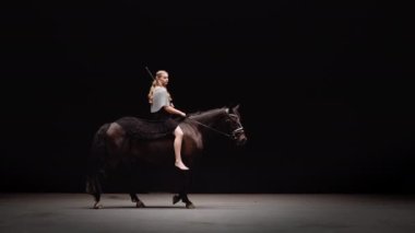 Genç Savaşçı Kadın Zincirli Giyinik ve Elinde Kılıçla Stüdyoda Siyah Arkaplan Karşısında At Biniyor. Horse Walks Around a Circle 'da. Yavaş Hareket Görüntüsü