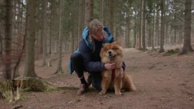Genç bir adam ormanda sevimli Chow Chow köpeğine sarılıp onu öperken çömeliyor.