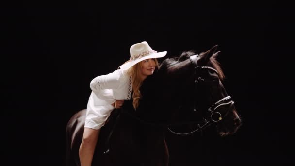 身穿白衣 头戴帽子的年轻女子骑着马 面对着黑色背景的摄影棚 她笑着躺在马背上 慢动作 多利射中 — 图库视频影像