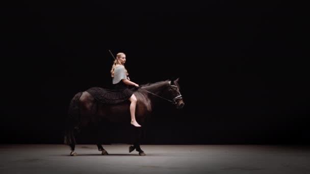 チェーンメールで身に着けていた若い女戦士と 黒を背景に馬に乗る彼女の手の剣 円の周りを馬が歩く スローモーションショット — ストック動画
