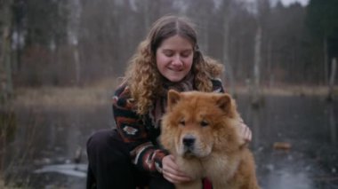 Sarışın bir kadın gülümseyip kameraya poz veriyor. Sevimli Chow Chow köpeğinin tüylerini okşayıp okşuyor.