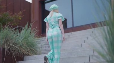 Sarışın bir kadının kareli bir akuamarin elbisesi ve örgü bir bereyle gündüz vakti bir moda çekiminde üst katta yürürken çekilmiş bir fotoğrafı.