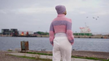 Sıcak pembe ve mor yün kazaklı bir kadın limana bakıyor.