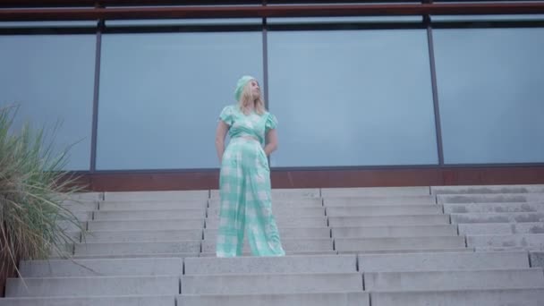 昼間の階段の飛行にポーズをとって チェックされたアクアマリンの服と編んだベレー帽を身に着けているブロンドの女性にズームワイドショット — ストック動画