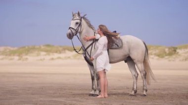 Beyaz elbiseli esmer bir kızın gündüz vakti kumlu bir toprakta atına sarılırken geniş bir fotoğrafı.