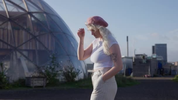 一名金发女郎身穿白衬衫 头戴粉色贝雷帽 白天双手插在口袋里 走在大地测量圆顶附近的一张中等全景照片 — 图库视频影像
